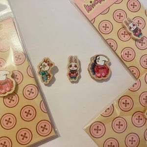May include: Small wooden buttons featuring colorful cartoon character illustrations. The set includes individual buttons and buttons attached to branded backing cards with a repeating pink button pattern and the text buttons knickknacks.