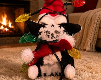 May include: A plush black and white cat toy wearing a red Santa hat with a white pom-pom. The cat is wrapped in a string of multicolored Christmas lights with red, yellow, and green bulbs. The toy has stitched facial features and white paws.