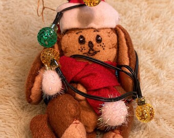 May include: A brown felt rabbit plush toy wearing a red Santa hat with a white trim and a red scarf. The rabbit is wrapped in a string of decorative Christmas lights featuring small, textured, multi-colored bulbs and fuzzy pink pom-poms.