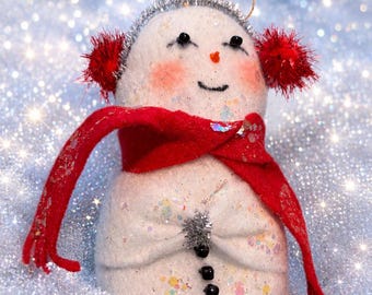 May include: A white felt snowman ornament featuring a bright red scarf, red fuzzy earmuffs, and a silver tinsel headband. The snowman has black bead eyes, a small orange nose, pink cheeks, and three black bead buttons down the front torso.