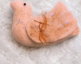 May include: A soft, pale pink felt bird ornament featuring a small black bead eye, gold glitter accents on the wing, and a thin, knotted copper-colored thread tied around the wing. The bird has a rounded body shape and a simple, stylized design.