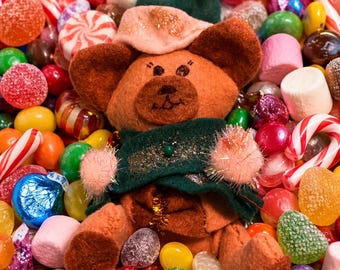 May include: A brown felt teddy bear ornament wearing a dark green cape and a matching pointed hat with a pink pom-pom. The bear has pink fuzzy cuffs, stitched facial features, and is surrounded by a colorful assortment of assorted holiday candies.