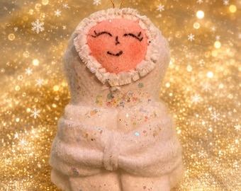 May include: A felt ornament shaped like a swaddled baby with a peach-colored face, embroidered closed eyes and mouth, and a white hood. The body is wrapped in white fabric covered in iridescent glitter, featuring a gold hanging loop at the top.