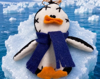 May include: A felt penguin ornament featuring a white face with black stitched X-shaped eyes, an orange beak, and a dark blue felt scarf. The penguin has black and white wings and orange feet, with a small gold string loop attached to the top.