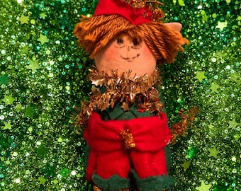 May include: A fabric elf doll featuring a green felt hat with a red pom-pom, brown yarn hair, and a hand-drawn face. The doll wears a green outfit with a red fabric belt, gold tinsel trim around the neck and shoes, and red pom-poms on the feet.
