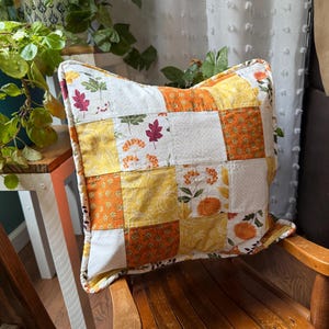 May include: A square throw pillow featuring a patchwork design of various fabrics in shades of orange, yellow, and white. The fabric patterns include autumn leaves, pumpkins, floral motifs, and small dots, creating a seasonal, multi-textured appearance.