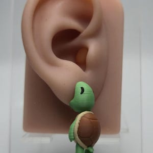 Pode incluir: Um pequeno brinco de tartaruga tridimensional com cabeça e membros verde-claros e uma carapaça castanha texturizada. A tartaruga está representada numa posição de pé, fixada num modelo de exposição em forma de orelha de cor bege.