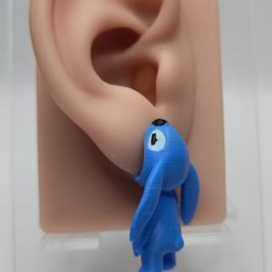 Pode incluir: Um brinco tridimensional de um personagem azul em forma de pequena criatura, com orelhas longas, nariz preto e manchas brancas nos olhos. A figura está posicionada verticalmente, parecendo pendurada no lóbulo da orelha com textura.
