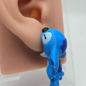 Pode incluir: Um brinco tridimensional azul com o formato da personagem Stitch, apresentando um rosto azul-claro, orelhas azul-escuras e detalhes pretos no nariz e nos olhos. O brinco está preso a um modelo de orelha de silicone bege realista.