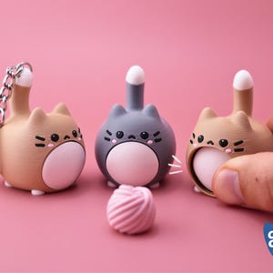 May include: Three cat-shaped figurines in tan and gray, with white tails and pink bellies, sit on a pink surface. One is attached to a keychain. A pink, swirled candy is in front of the gray cat. The figurines have black facial details.