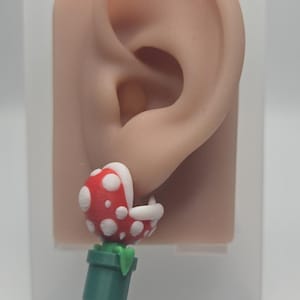 Pode incluir: Um brinco de planta piranha vermelha com pontos brancos e uma base de vaso verde. A planta tem a boca aberta com dentes brancos visíveis e está presa a um vaso cilíndrico verde. O design apresenta um acabamento fosco.