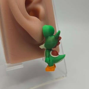 Pode incluir: Um brinco em forma de dinossauro verde com uma sela vermelha, crista vermelha e pés cor de laranja. A personagem está representada de pé, com um focinho branco e cabeça arredondada, exibindo uma textura típica de impressão 3D.
