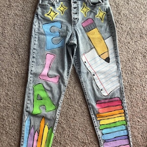 May include: Light gray acid-wash denim jeans featuring hand-painted school-themed designs. The left leg displays the name ELA in large colorful letters above a row of crayons. The right leg shows a pencil, lined paper, and a stack of books.