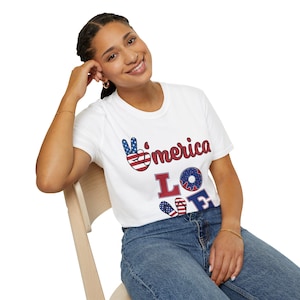 Puede incluir: Una camiseta blanca de manga corta con un diseño gráfico que incluye el texto "merica" y "LOVE". La letra "A" se sustituye por un signo de la paz, la "O" es un dónut y la "V" es un par de chanclas, todo en rojo, blanco y azul.
