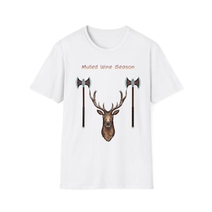 Puede incluir: Camiseta blanca con una cabeza de ciervo y dos hachas. El texto "Mulled Wine Season" está encima del ciervo. Las hachas tienen hojas plateadas y mangos marrones oscuros. El ciervo es marrón con grandes cuernos.