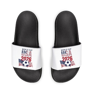 Puede incluir: Un par de sandalias deslizantes negras con correas blancas. Las correas presentan el texto "WORLD CUP USA 2026" en rojo y azul, junto con un balón de fútbol y un diseño de la bandera estadounidense.