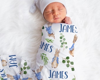 May include: A white swaddle blanket with a repeating pattern of brown rabbits wearing blue jackets and green leaves. The blanket is wrapped around a sleeping baby wearing a light blue hat. The name "JAMES" is printed on the blanket in blue.