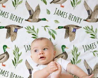 May include: A white baby blanket with a repeating pattern of green leaves and watercolor ducks. The blanket features the name "James Henry" repeated throughout the design.