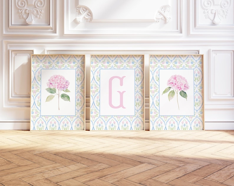 May include: A set of three rectangular wall art prints featuring a light blue and pink floral trellis pattern border. The two outer prints display a single pink hydrangea flower with green leaves, while the center print features a large pink letter G.