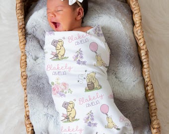 May include: A newborn baby wrapped in a personalized blanket with a Winnie the Pooh theme, nestled in a woven basket. The blanket features illustrations of Pooh Bear, flowers, and the name "Blakely Amelia". A white bow adorns the baby's head.