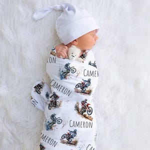 May include: White swaddle blanket with a motocross theme. The blanket features multiple images of dirt bikes and the name "Cameron" repeated throughout.