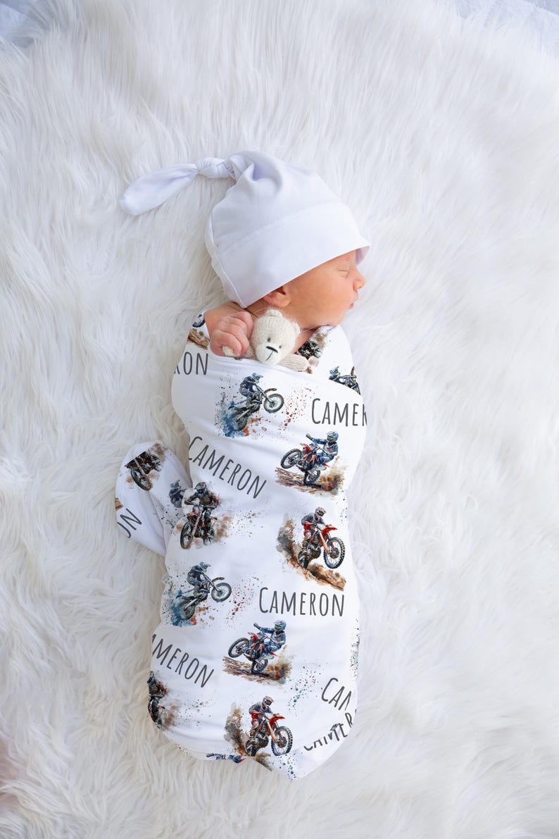 May include: White swaddle blanket with a motocross theme. The blanket features multiple images of dirt bikes and the name "Cameron" repeated throughout.