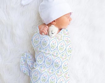 May include: A newborn baby wearing a white knotted cap and wrapped in a patterned swaddle. The swaddle features a blue and pink floral design. A small white teddy bear is held near the baby's face, all resting on a white fluffy surface.