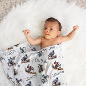 May include: A white baby blanket with a watercolor design of a person riding a dirt bike. The blanket is covered in the name "Cameron" repeated multiple times.