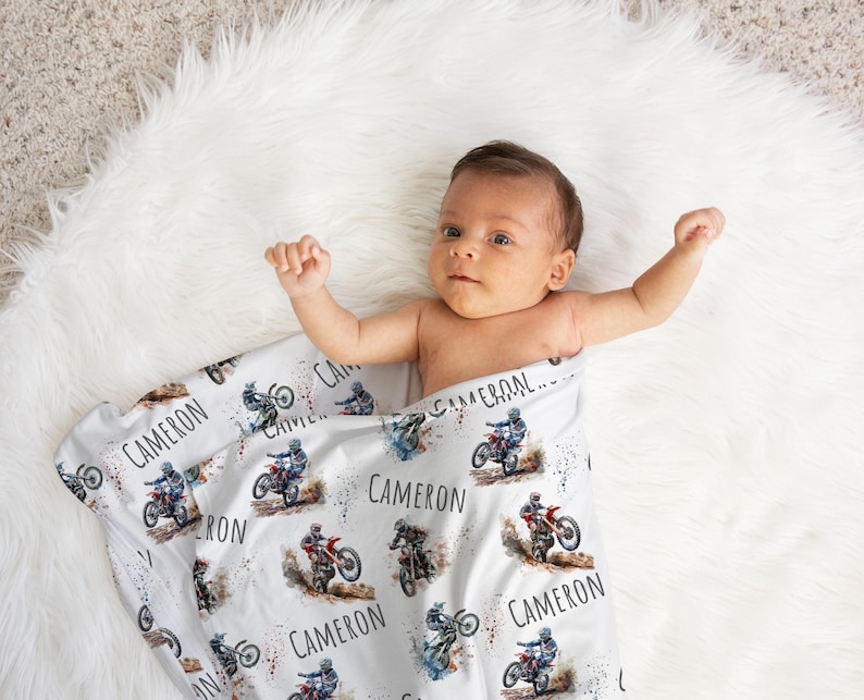 May include: A white baby blanket with a watercolor design of a person riding a dirt bike. The blanket is covered in the name "Cameron" repeated multiple times.