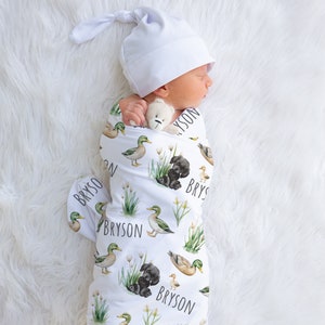 May include: A white swaddle blanket with a pattern of ducks, puppies, and green grass. The blanket has the name "Bryson" printed on it multiple times. A baby is sleeping in the swaddle, holding a stuffed animal.