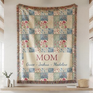 May include: A woven throw blanket with a patchwork design featuring floral and watercolor patterns in shades of blue, pink, and cream. The word "MOM" is displayed prominently, with the names "Grace - Joshua - Madeline" below.
