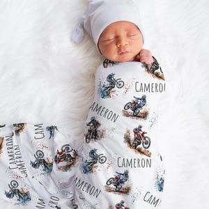May include: A white swaddle blanket with a watercolor design of motocross riders. The blanket features the name "Cameron" repeated throughout the design.