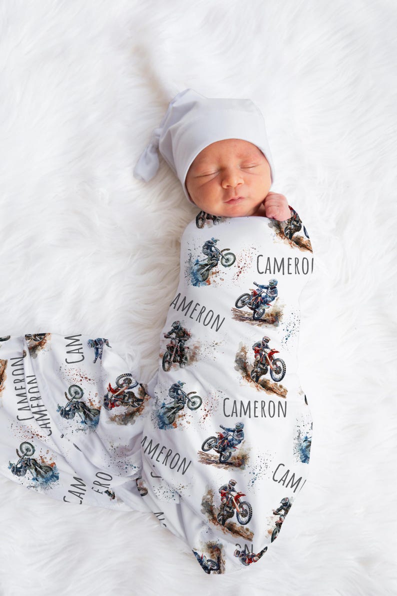 May include: A white swaddle blanket with a watercolor design of motocross riders. The blanket features the name "Cameron" repeated throughout the design.
