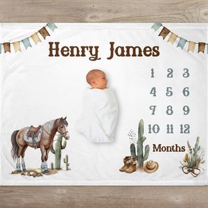 May include: A white baby blanket with a brown horse illustration, cactus, and cowboy boots. The blanket has a banner with the name "Henry James" and a monthly growth chart with numbers 1-12.