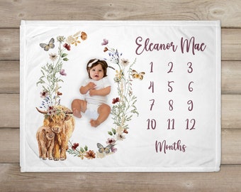 May include: White baby blanket with a floral wreath design, a baby girl, and two Highland cows. The blanket has a pink and white color scheme and includes the text "Eleanor Mae" and a number chart for tracking a baby's growth from 1 to 12 months.