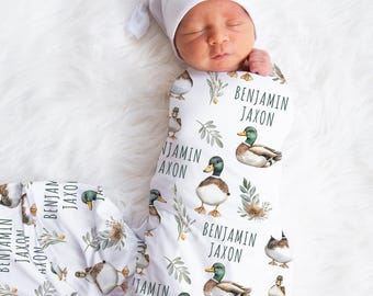 May include: A newborn baby swaddled in a white blanket and matching cap. The blanket features a pattern of ducks, greenery, and the name "BENJAMIN JAXON" in black text. The baby is lying on a fluffy white surface.