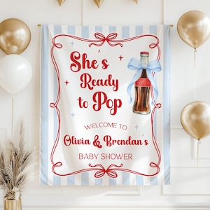 May include: A baby shower banner with the text "She's Ready to Pop" in red script. The banner features a cartoon illustration of a soda bottle with a blue ribbon. The background has light blue vertical stripes and red decorative borders.