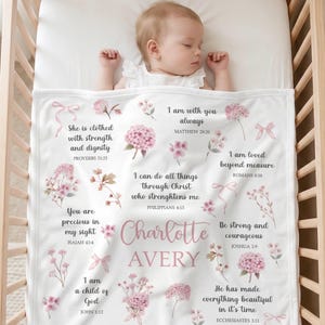 May include: A white baby blanket featuring the name Charlotte Avery in pink script and block lettering, surrounded by watercolor illustrations of pink flowers, ribbons, and various Bible verses printed in a dark gray serif font.