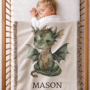 May include: A beige baby blanket with a green dragon illustration and the name "MASON" in black. The blanket has a fringed edge and is in a wooden crib. A baby is sleeping on the blanket.
