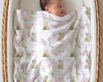 May include: A white baby blanket with a repeating pattern of a yellow bear, pink flowers, and pink balloons. The name "Emersyn Clare" is printed in pink. The blanket is in a wicker bassinet.