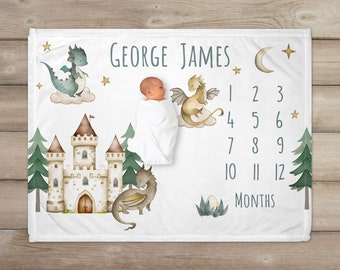 May include: A white baby blanket with a castle, dragons, stars, and a moon. The blanket has a number chart for tracking a baby's growth from 1 to 12 months. The text "GEORGE JAMES" is printed on the blanket.