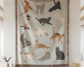 May include: A light blue throw blanket featuring illustrations of various cat breeds in different poses. The blanket has a fringed edge with a mix of colors. The cats are in shades of brown, gray, black, and white.