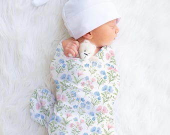 May include: A newborn baby swaddled in a floral print blanket, wearing a white cap, and clutching a small teddy bear. The blanket features a pattern of blue and pink flowers on a white background. The baby is lying on a fluffy white surface.
