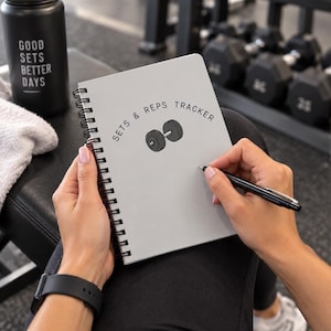 May include: A light gray spiral-bound notebook with the text "SETS & REPS TRACKER" and a black dumbbell illustration on the cover. The notebook is held open with a black pen, and a black water bottle with the text "GOOD SETS BETTER DAYS" is nearby.