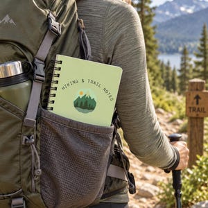 Könnte beinhalten: Ein hellgrünes Notizbuch mit Spiralbindung, dem Text HIKING & TRAIL NOTES und einer Grafik aus Berg, Wald und Sonne. Das Notizbuch steckt in der Seitentasche eines olivgrünen Rucksacks, der auch eine metallische Wasserflasche enthält.