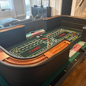 May include: A large, U-shaped craps table with a green felt playing surface. The table has wooden rails and black sides. Stacks of colorful casino chips are arranged around the table. The word "COME" is prominently displayed on the felt.