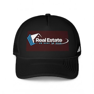 May include: A black trucker hat featuring a rectangular graphic on the front panel. The graphic displays a white house icon with blue accents, the text Real Estate, and the text I AM HERE TO HELP in blue, set against a dark maroon background.