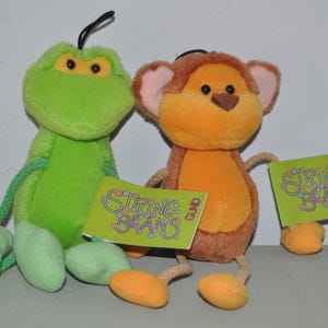 May include: Two Gund String Beans plush toys, one green frog and one brown monkey, each featuring long rope-like limbs, soft fabric bodies, and attached lime green rectangular tags with the text String Beans printed in purple and blue lettering.