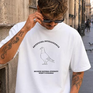 May include: A white short-sleeved t-shirt featuring a black line drawing of a pigeon in the center. Above the bird, the text reads ARTIFICIAL INTELLIGENCE, and below the bird, the text reads BECAUSE NATURAL STUPIDITY WASN'T SCALABLE.