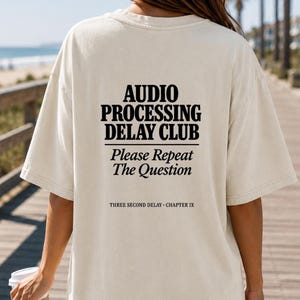 May include: Cream-colored t-shirt with the text "AUDIO PROCESSING DELAY CLUB" in black, with "Please Repeat The Question" below. The phrase "THREE SECOND DELAY - CHAPTER IX" is printed at the bottom.
